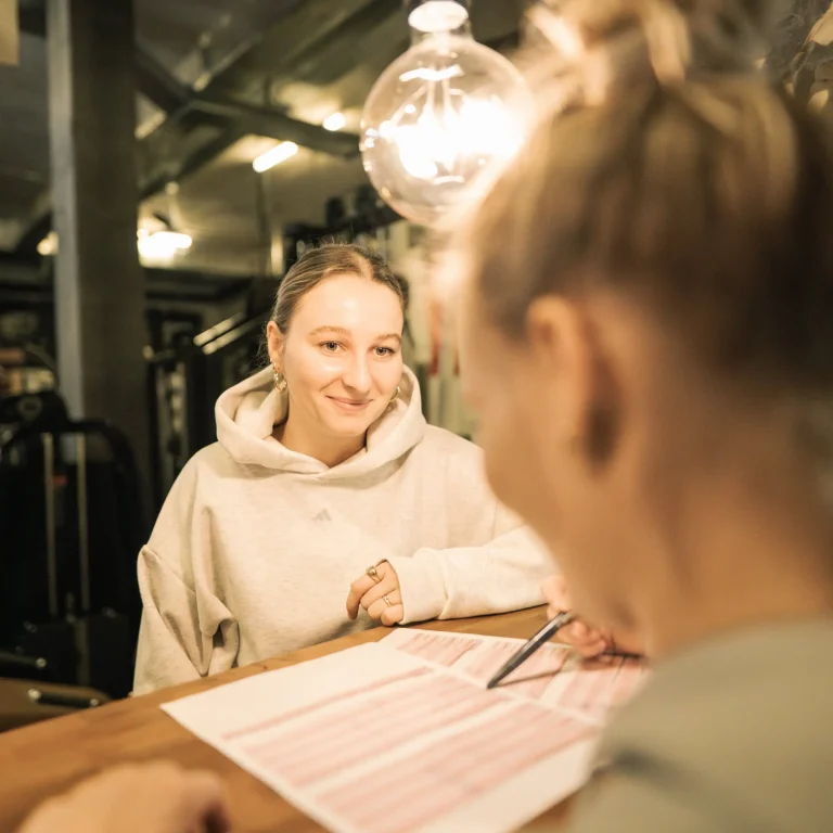 Zwei Frauen sitzen sich an der Bar von Legym gegenüber, während eine junge Frau in einem grauen Hoodie aufmerksam zuhört. Auf dem Tisch liegt ein Ernährungsplan von Legym, auf den die andere Frau mit einem Stift zeigt.