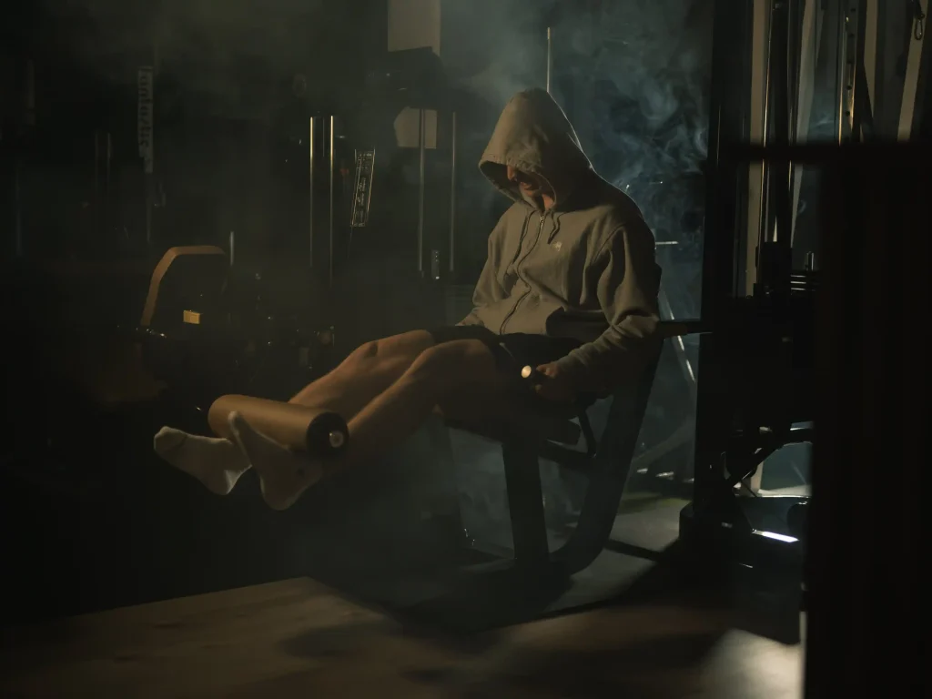 Junger Mann mit Kapuze, die an einem Fitnessgerät in einer düsteren, leicht nebligen Atmosphäre trainiert.